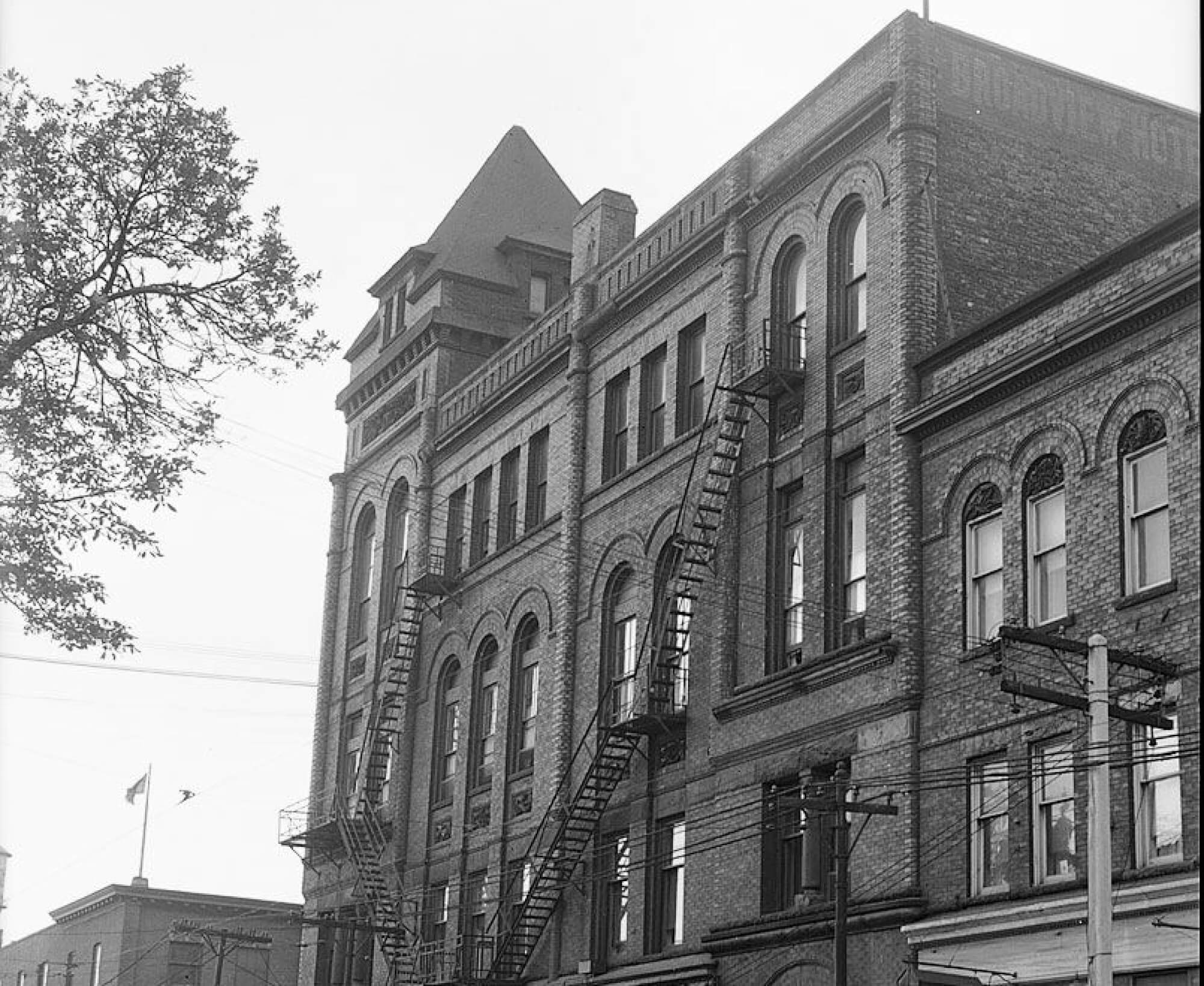 Black and white photo of a 4-storey building on a street corner. Old 1940s style cars are parked on the street beside the building.