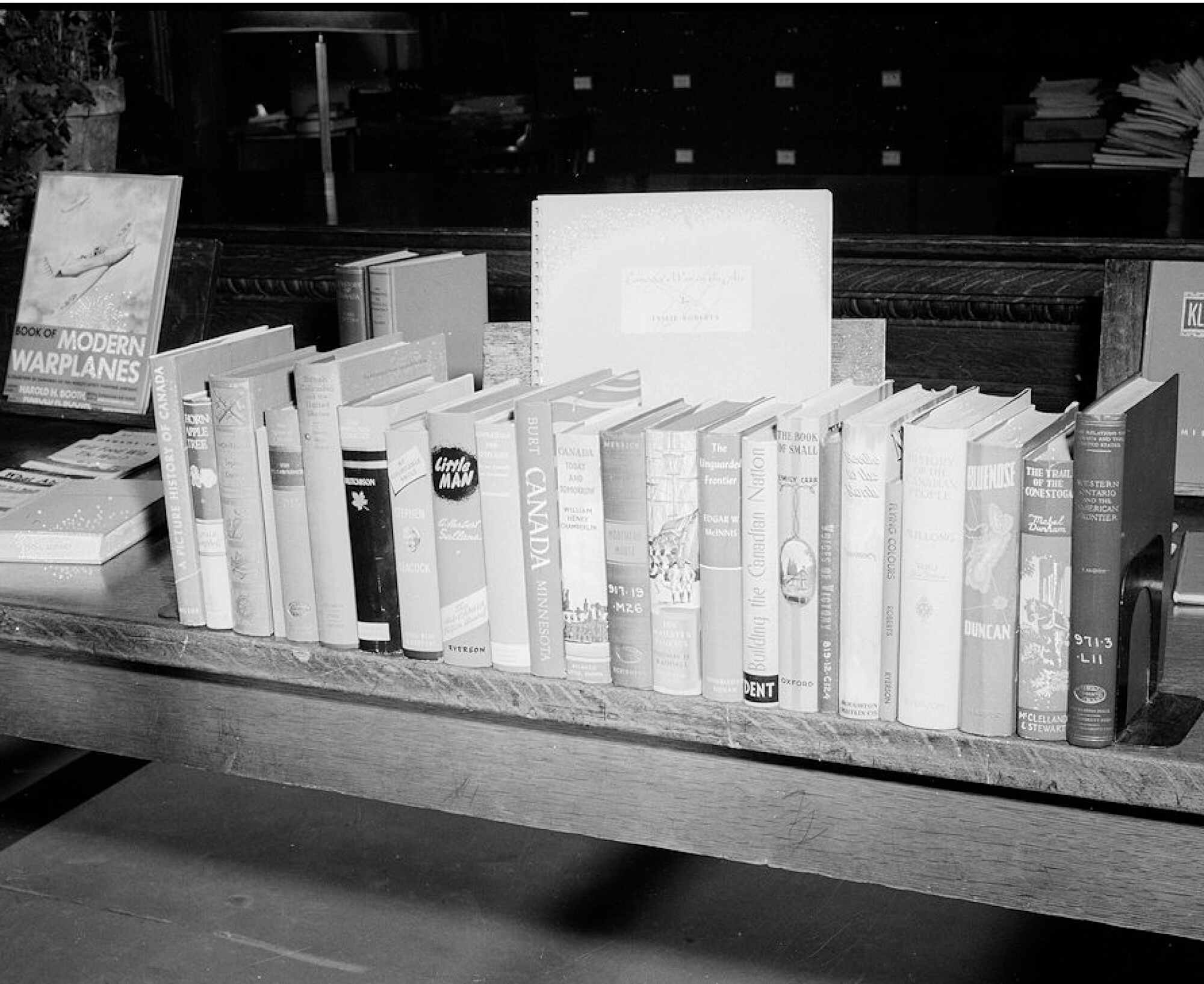 A black and white photograph of a row of books standing upright on a wooden table.