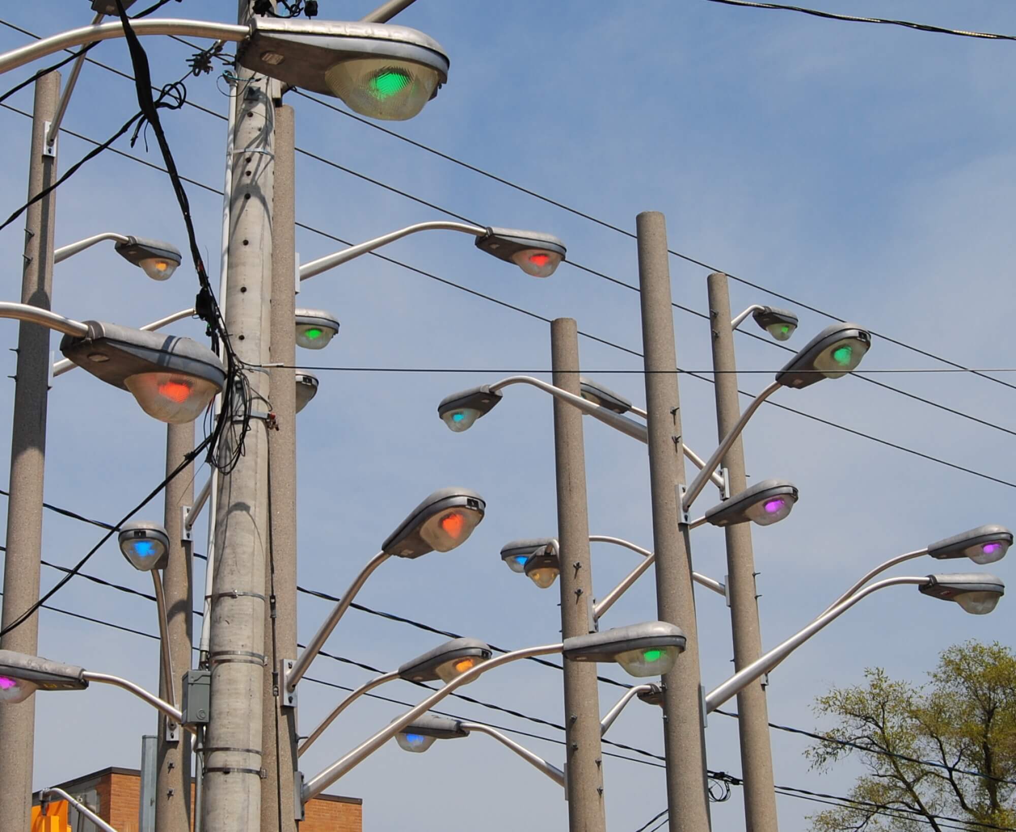 Art installation featuring eight light poles with several streetlights jutting out in different directions. The lights are a variety of colours: green, red, blue, orange, purple. Above are several electrical wires and a blue sky.
