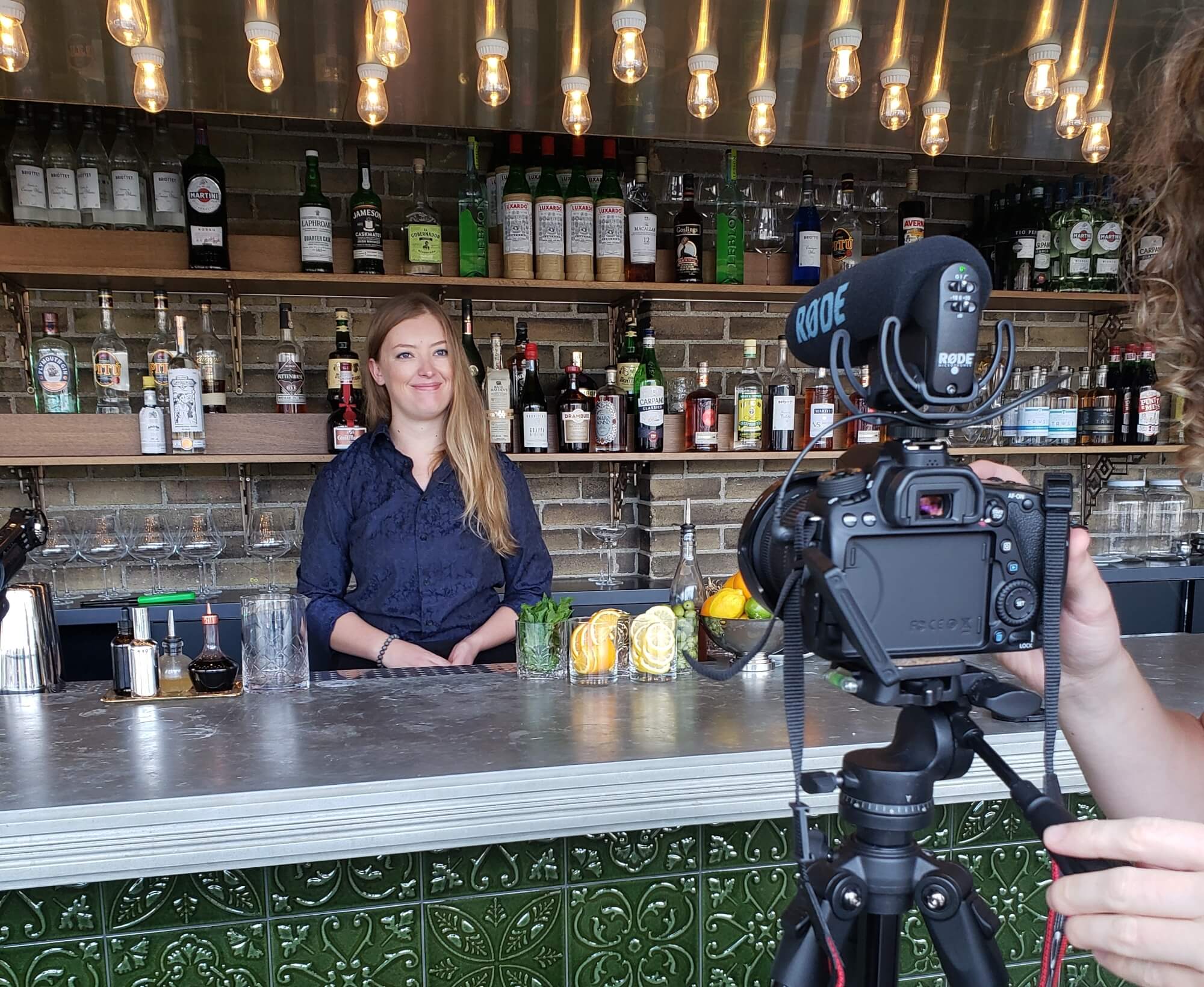 A women wearing a blue shirt stands behind a cocktail bar, smiling at a camera in the foreground of the image.