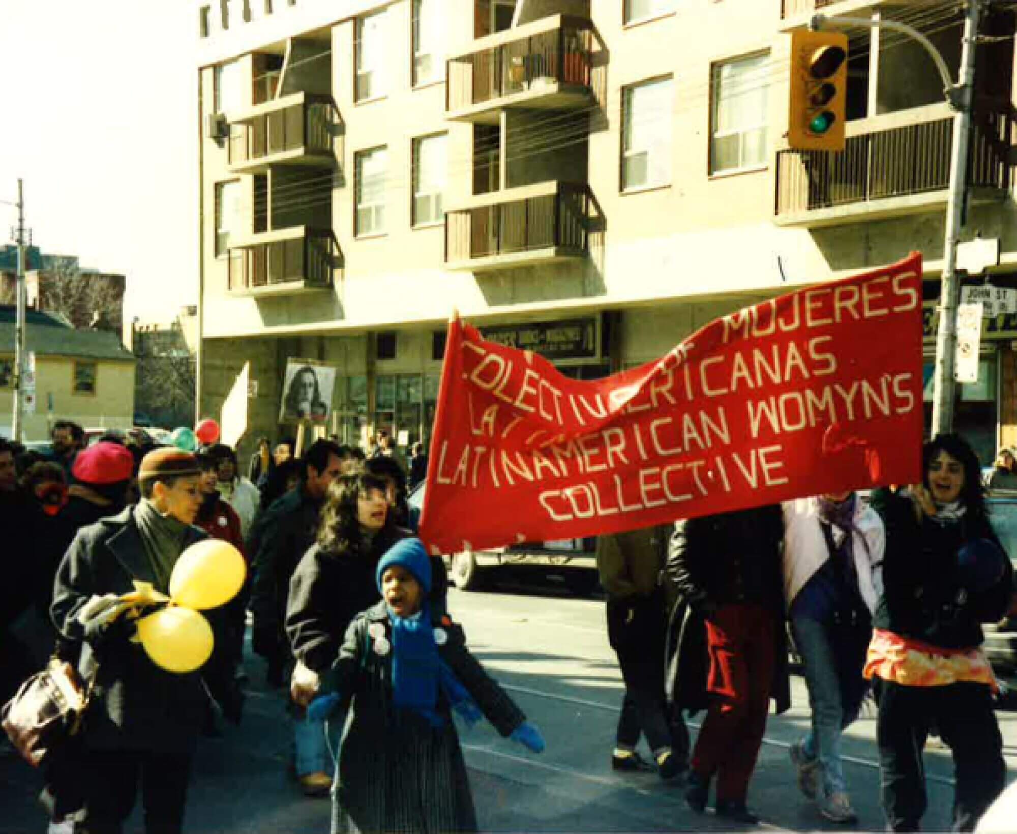 A group of women march on a city street in daylight. Two of them are holding a red banner with white lettering. The top of the banner is crumpled and much of the text is obscured. The word mujeres can be seen at the top, at the bottom the words Latin American Womyn's Collective can be seen. Above the group is single traffic light hanging from a street pole. Behind the group is the bottom half of a tall, beige apartment building.