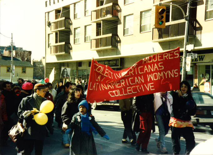 A group of women march on a city street in daylight. Two of them are holding a red banner with white lettering. The top of the banner is crumpled and much of the text is obscured. The word mujeres can be seen at the top, at the bottom the words Latin American Womyn's Collective can be seen. Above the group is single traffic light hanging from a street pole. Behind the group is the bottom half of a tall, beige apartment building.