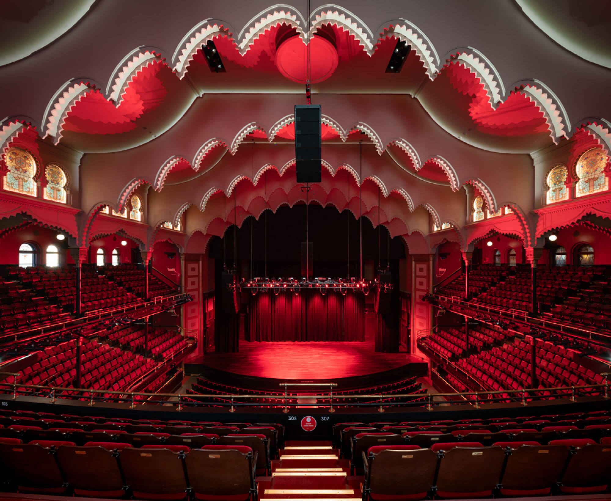 The interior of a music hall. The seating in the hall has red upholstery. On the ceiling of the hall are decorative archways that line up with the decorations on the stage in the center.