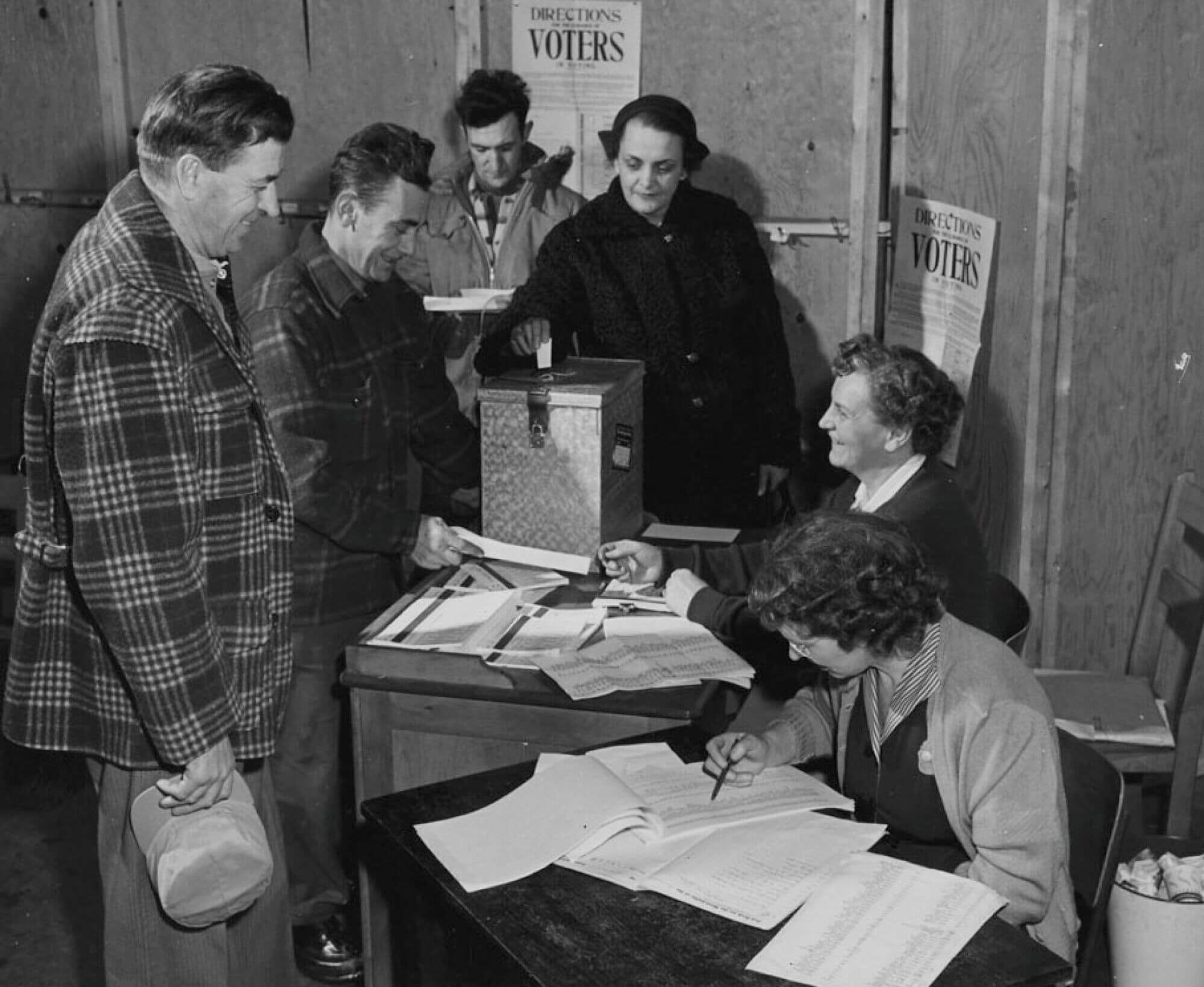 a black and white image of a woman seated at a voting booth. To the left are a group of voters ready to cast their vote. Another woman stands over a ballot box placing her ballot inside.