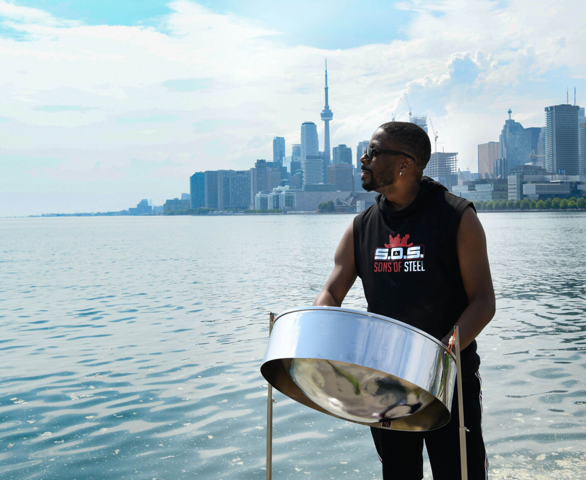 A man stands behind a steelpan. He wears a black sleeve-less shirt that reads "Sons of Steel." Behind him is a large body of water, behind which can be seen the city skyline of Toronto.