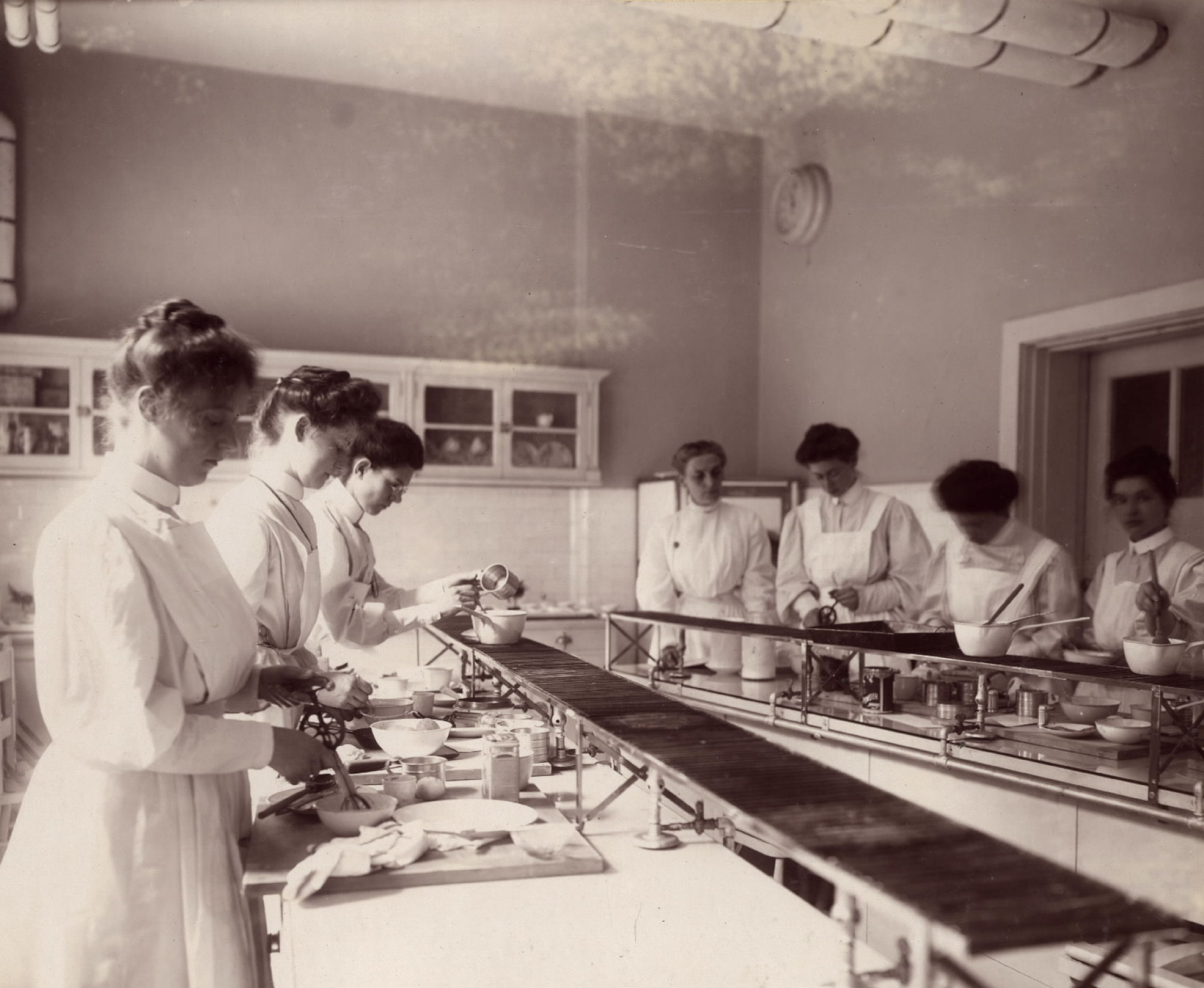 Sepia-toned image of nurses working in a kitchen plating food on long counters.
