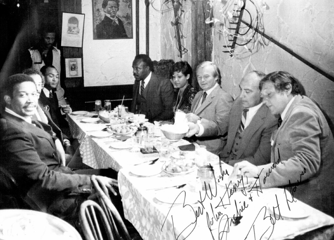 Photograph of a group of eight men in suits seated at a long restaurant table. The picture is signed "Best wishes John Henry, Archie, Howard, Best wishes, Bill Davis".
