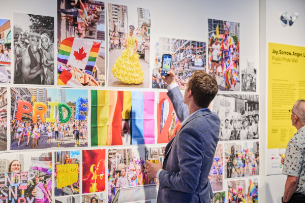 A man stands in front of a wall of colourful photos. He holds his phone up to take a photograph of the wall.