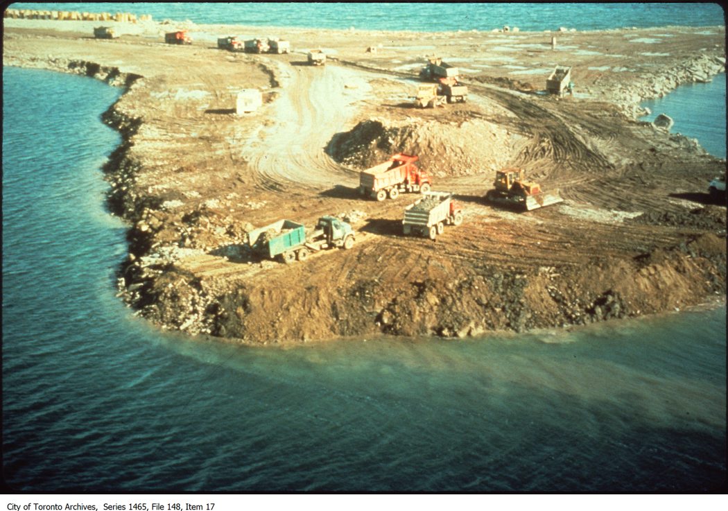 Colour photograph looking down on a rocky, brown strip of land, which takes up most of the image. Surrounding it is dark blue water, except for a strip of land at the top-left of the picture. A dirt trail leads from the top-left strip down to the centre of the landmass. Several large dump trucks move to and from a central mount of dirt. Another yellow construction vehicle is nearby. One of the dump trucks, painted blue, appears to be offloading soil close to the water's edge. 