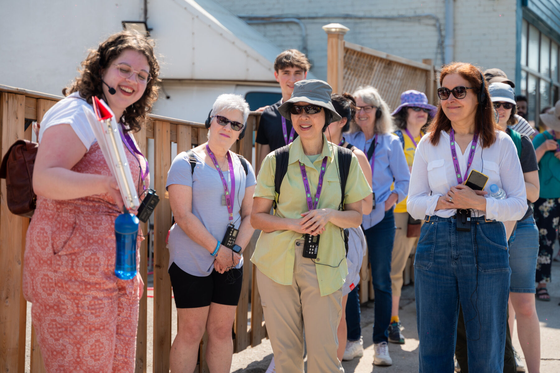 A person stands with a group of people on a sidewalk. She holds a binder open, and looks over at it with a smile on her face. People in the foreground look at the material being displayed.