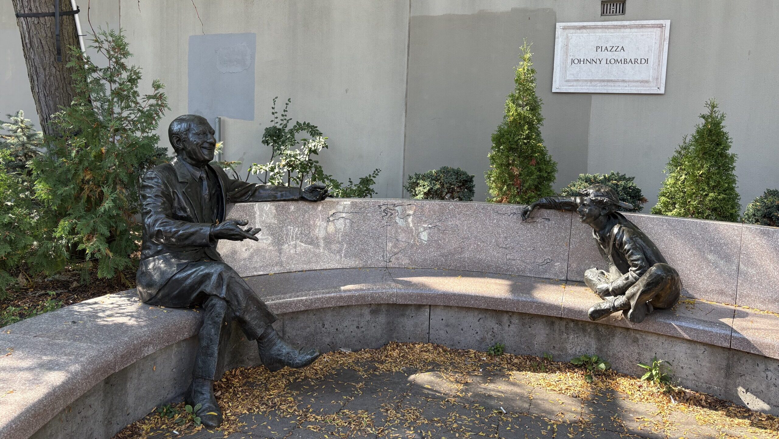 A bench in a park with a statue of a cheerful man and a young child sitting on it. A plaque can be seen above it reading "Piazza Johnny Lombardi".