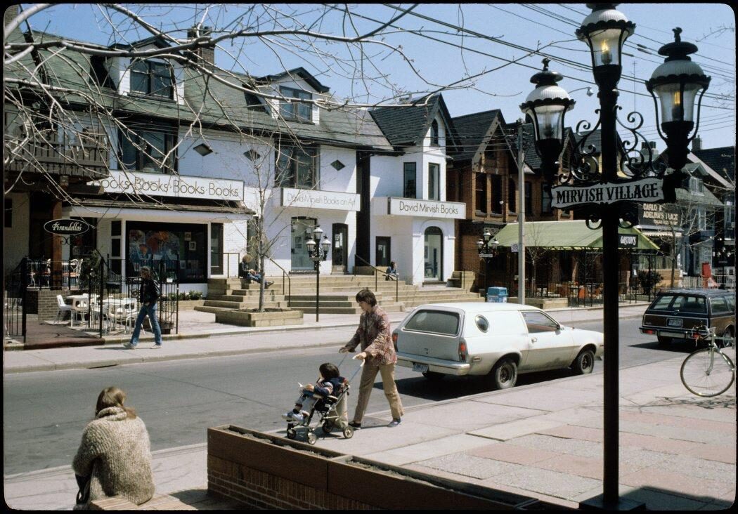 A street view of Mirvish Village on Markham Street, showing the David Mirvish Books storefront, people walking, a person pushing a stroller, and parked cars.