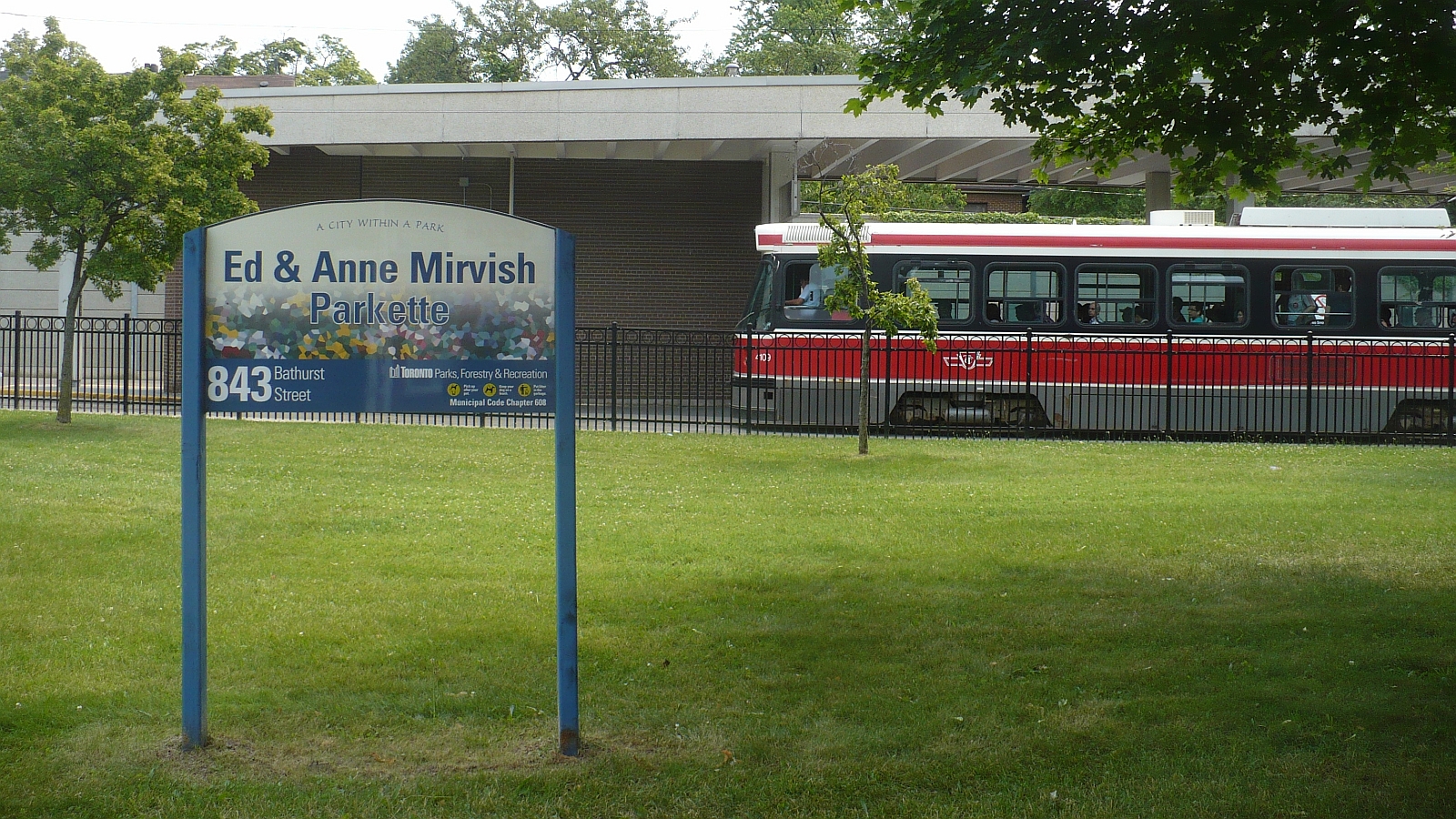 A lawn with a sign reading "Ed & Anne Mirvish Parkette." A streetcar with a red and white colour scheme is visible in the background, behind a fence.