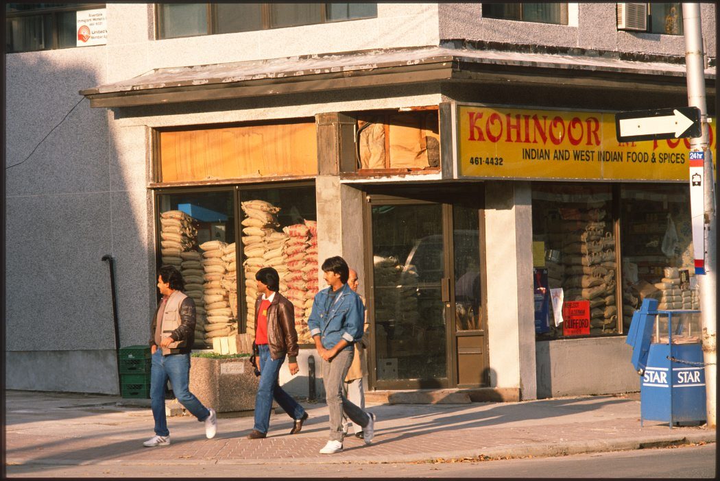 Three young South Asian men walking past a shop with a sign that reads Kohinoor Foods Indian and West Indian Food and Spices.