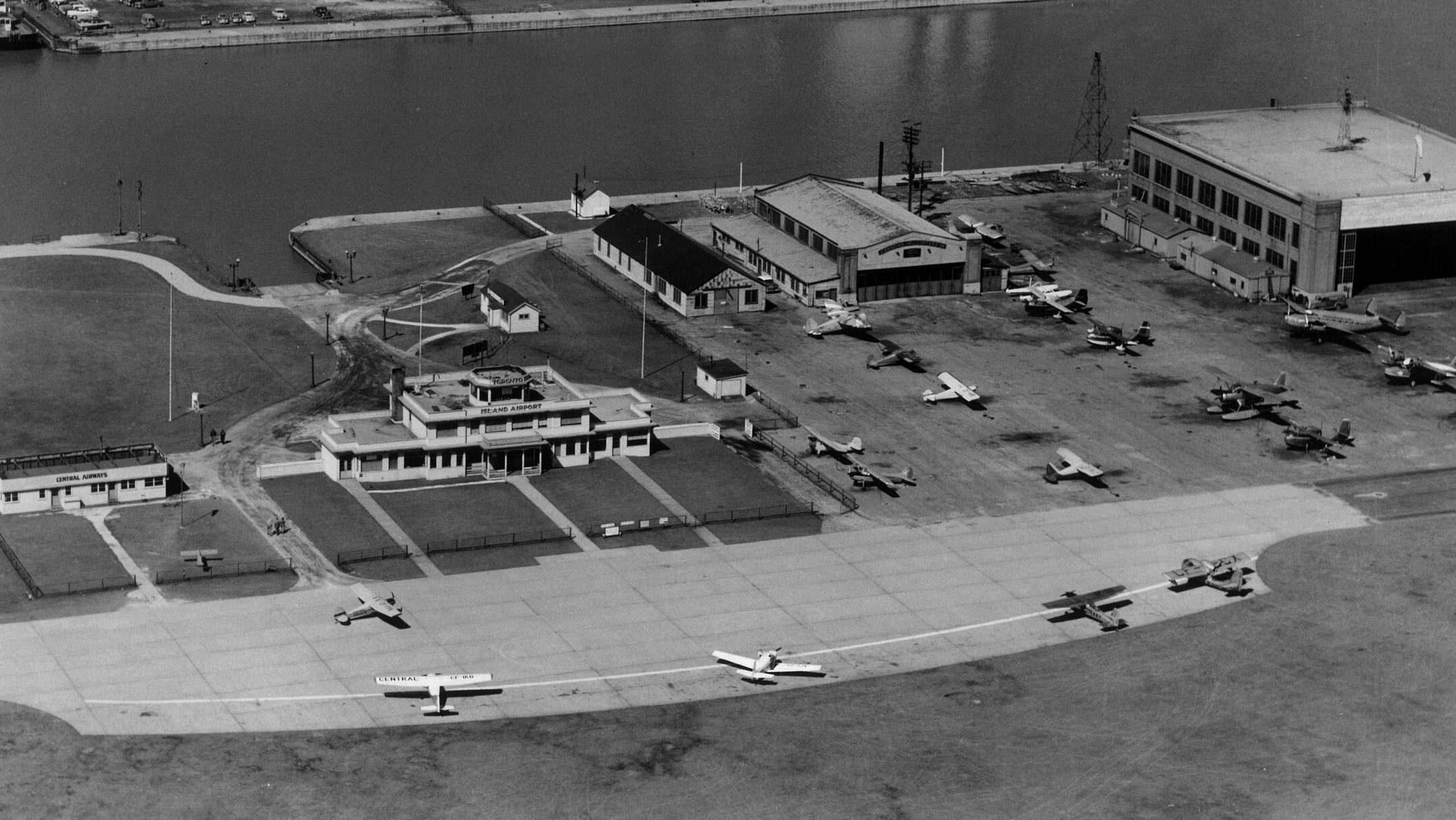 Black and white aerial view of the island airport in Toronto in 1957. An early style airport terminal sits at the base of the Western Gap next to three other structures, surrounded by many aircraft.