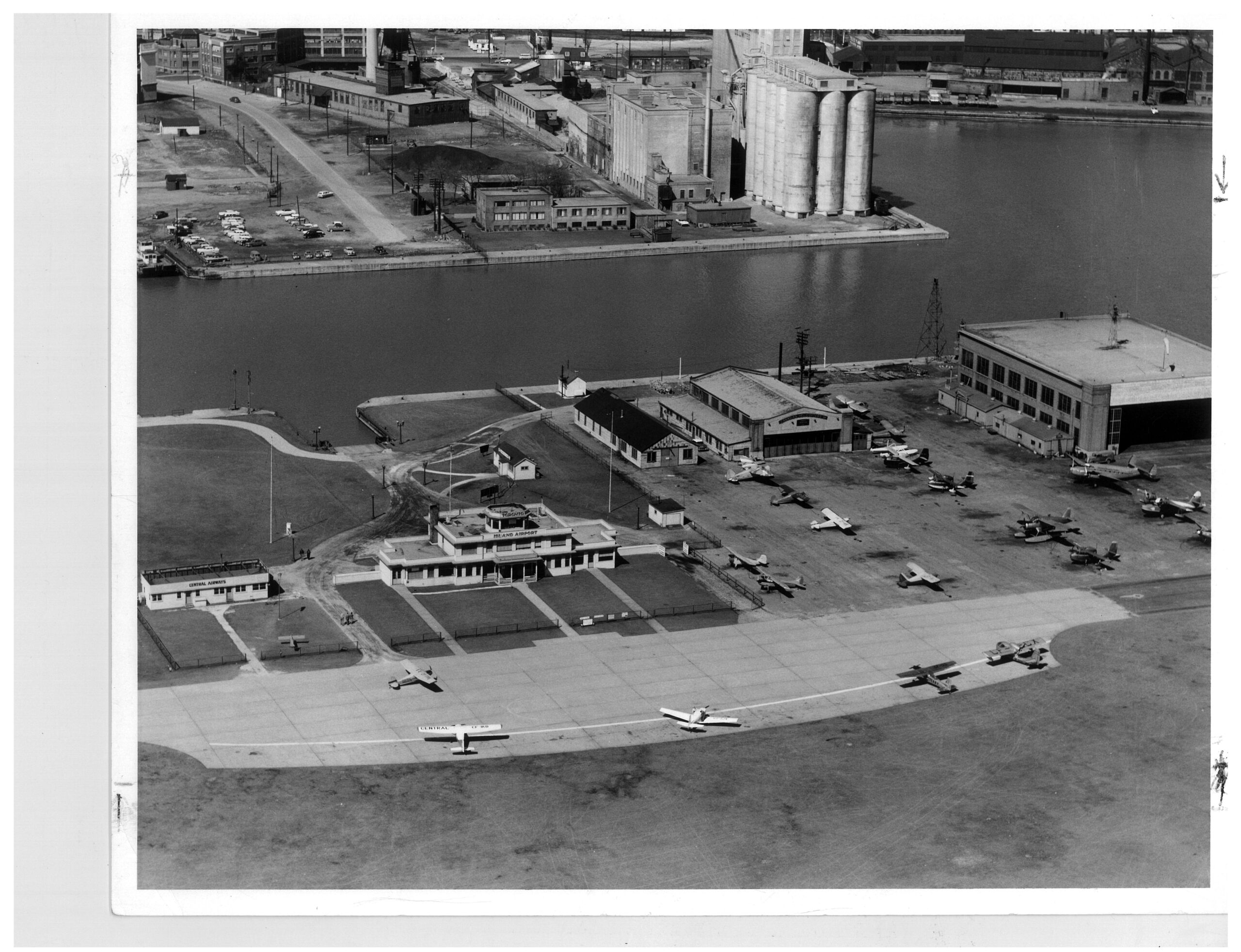Black and white aerial view of the island airport in Toronto in 1957. An early style airport terminal sits at the base of the Western Gap next to three other structures, surrounded by many aircraft.