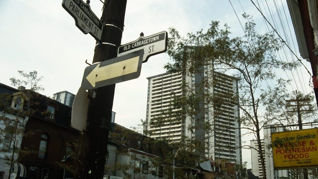 A coloured image of a street line with cars and businesses. High-rise apartment buildings are visible in the distance.