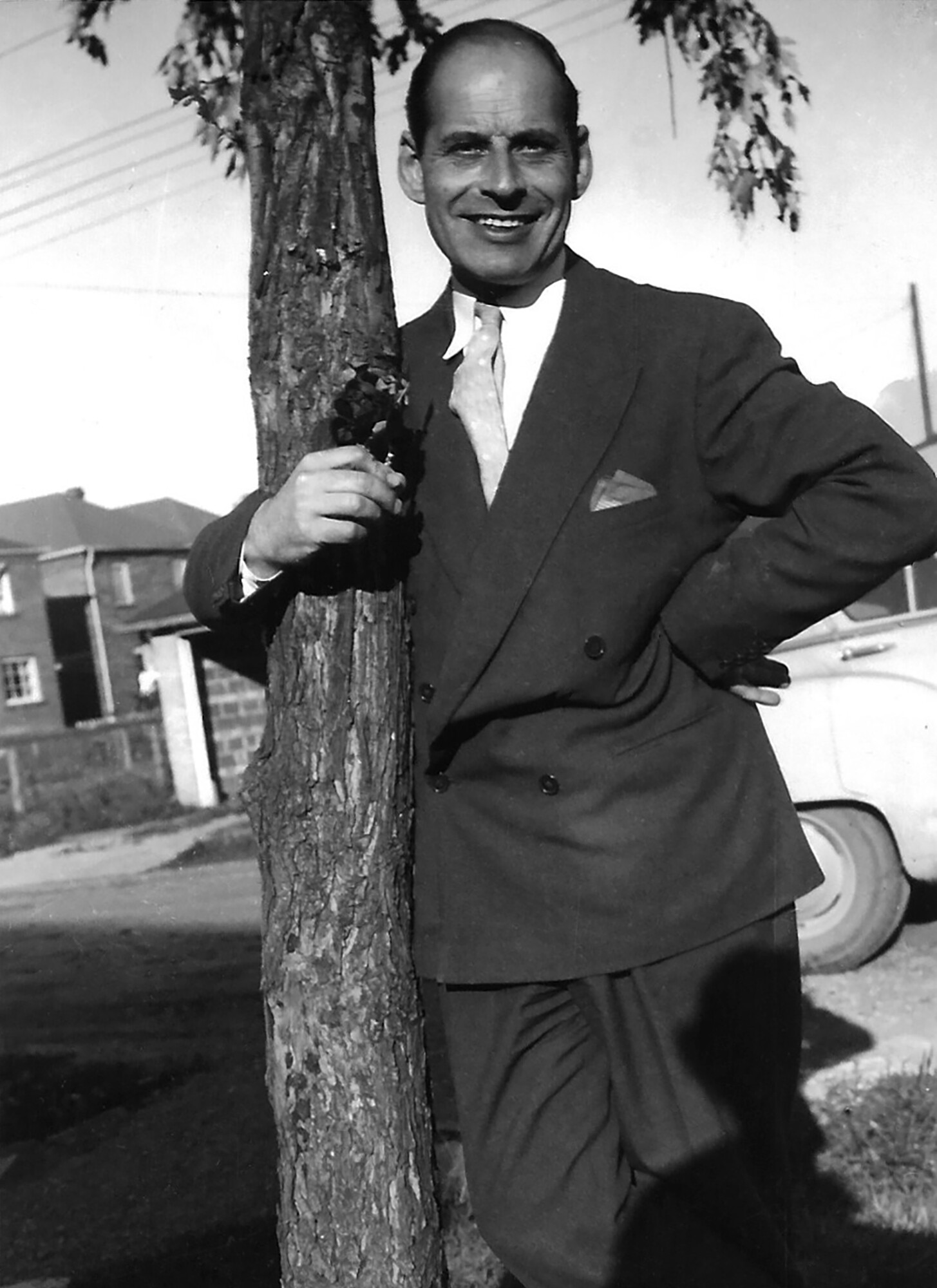 Image of a smiling man in a suit with his arm around a small tree. Part of a car and houses are visible in the background.