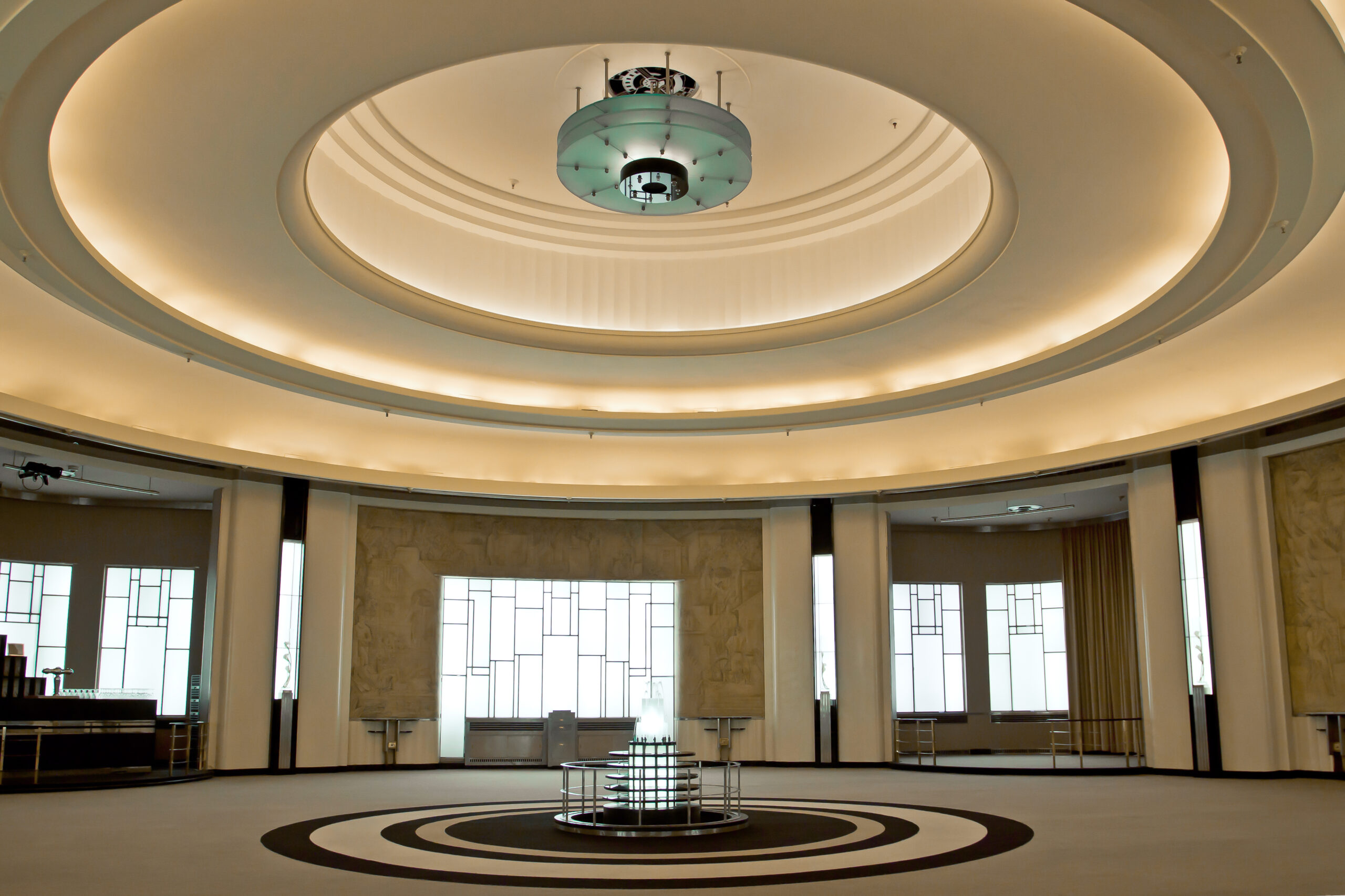 A brightly lit empty round room. The floor is covered with carpet and has a small fountain at its center. The ceiling features round sculptural elements and soft lighting.