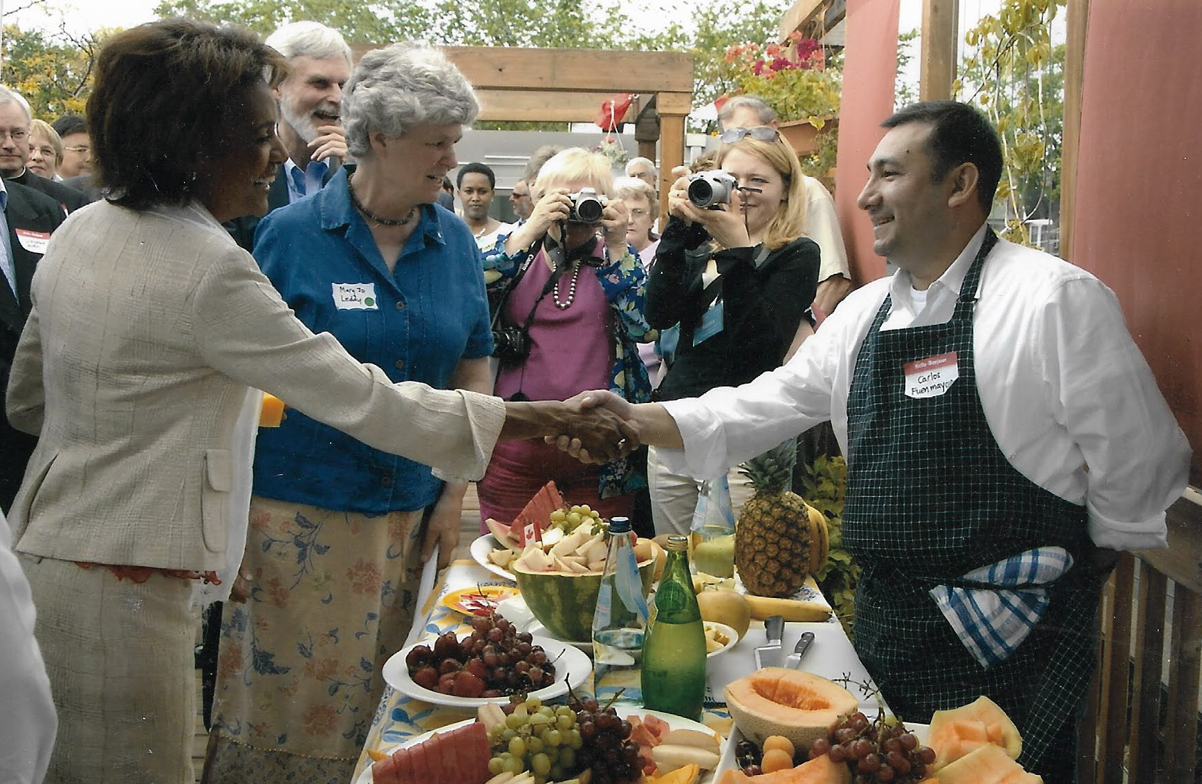 A crowd on a rooftop watches as a woman in a dress suit shakes hands with a man in an apron and chef's jacket. Two people are pointing cameras in their direction. In front of them is a table with various fruits cut up and on plates along with glass water bottles.