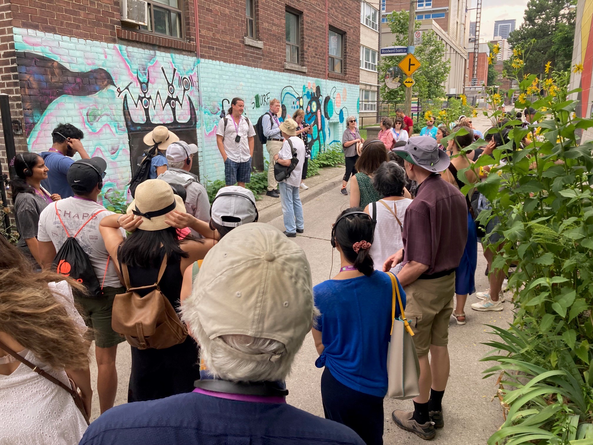 Group of people stand in an alley. On one side is a colourful mural on a brick wall. The other side has a planting of sunflowers.