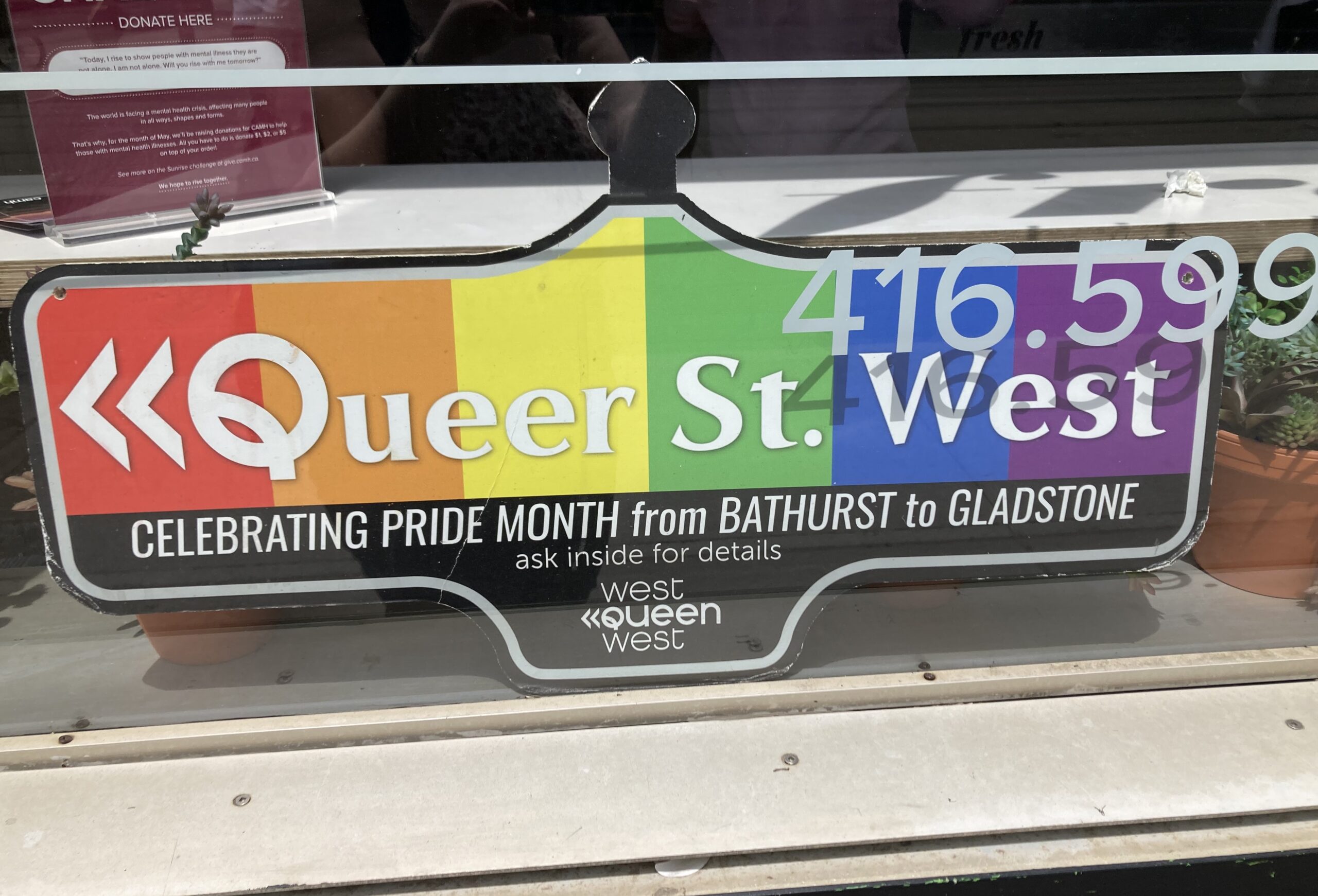 A sign shaped like an old-fashioned street sign with rainbow background sits on a counter behind a glass window. The sign reads “Queer St. West. Celebrating pride month from Bathurst to Gladstone. Ask inside for details. West Queen West.”
