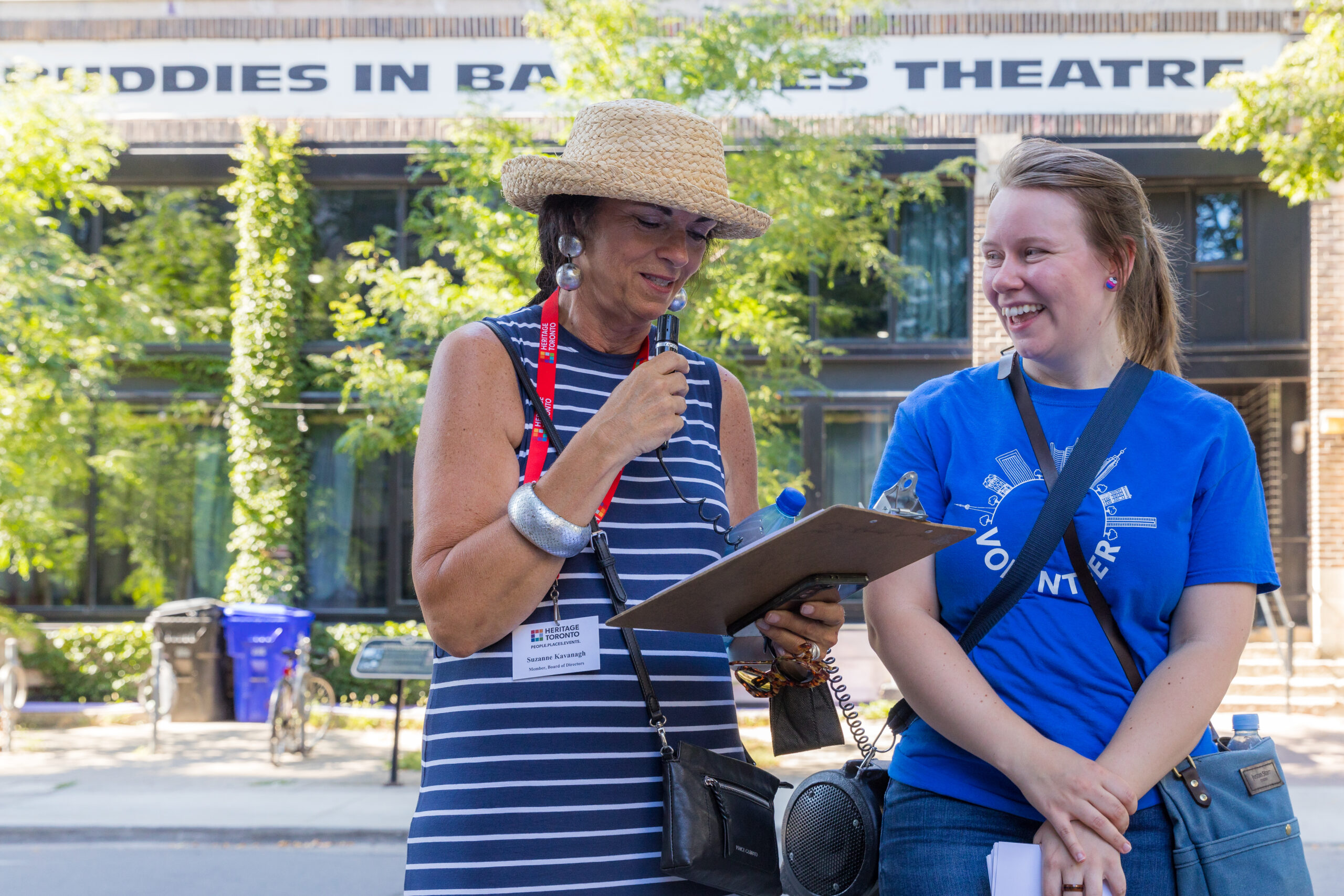 Two women stand beside each other. On the left, one holds a microphone and reads from a clipboard. On the right, one smiles at her companion.