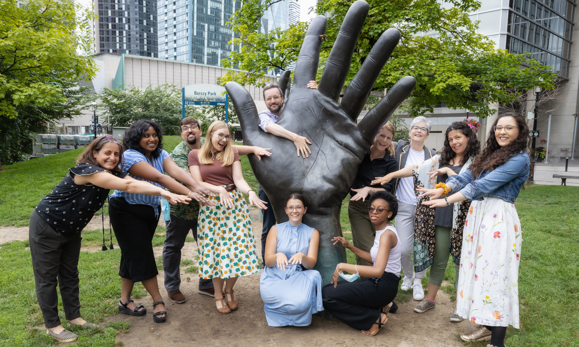Group of 11 people hold out their hands as they pose around a large hand sculpture in a park.