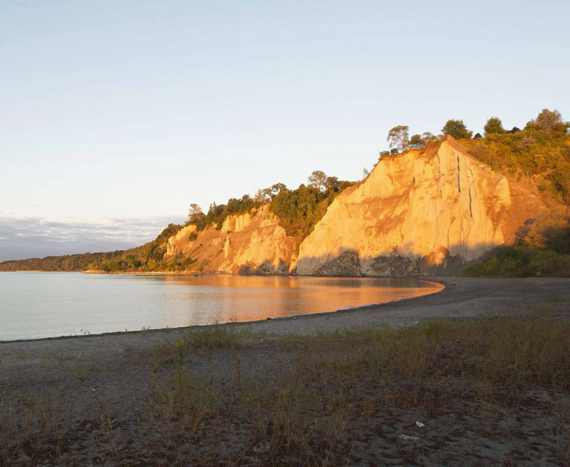 A scenic shoreline with a beach in the foreground. In the background, a natural bluff can be seen. It is sunset.