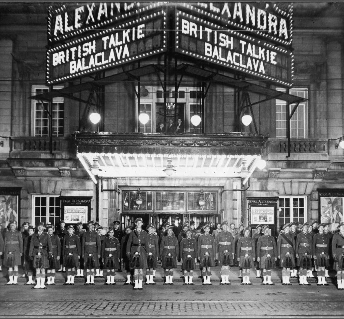 A black and white photo of a bright and lit up theatre. Rows of men in uniform stand in front of it.