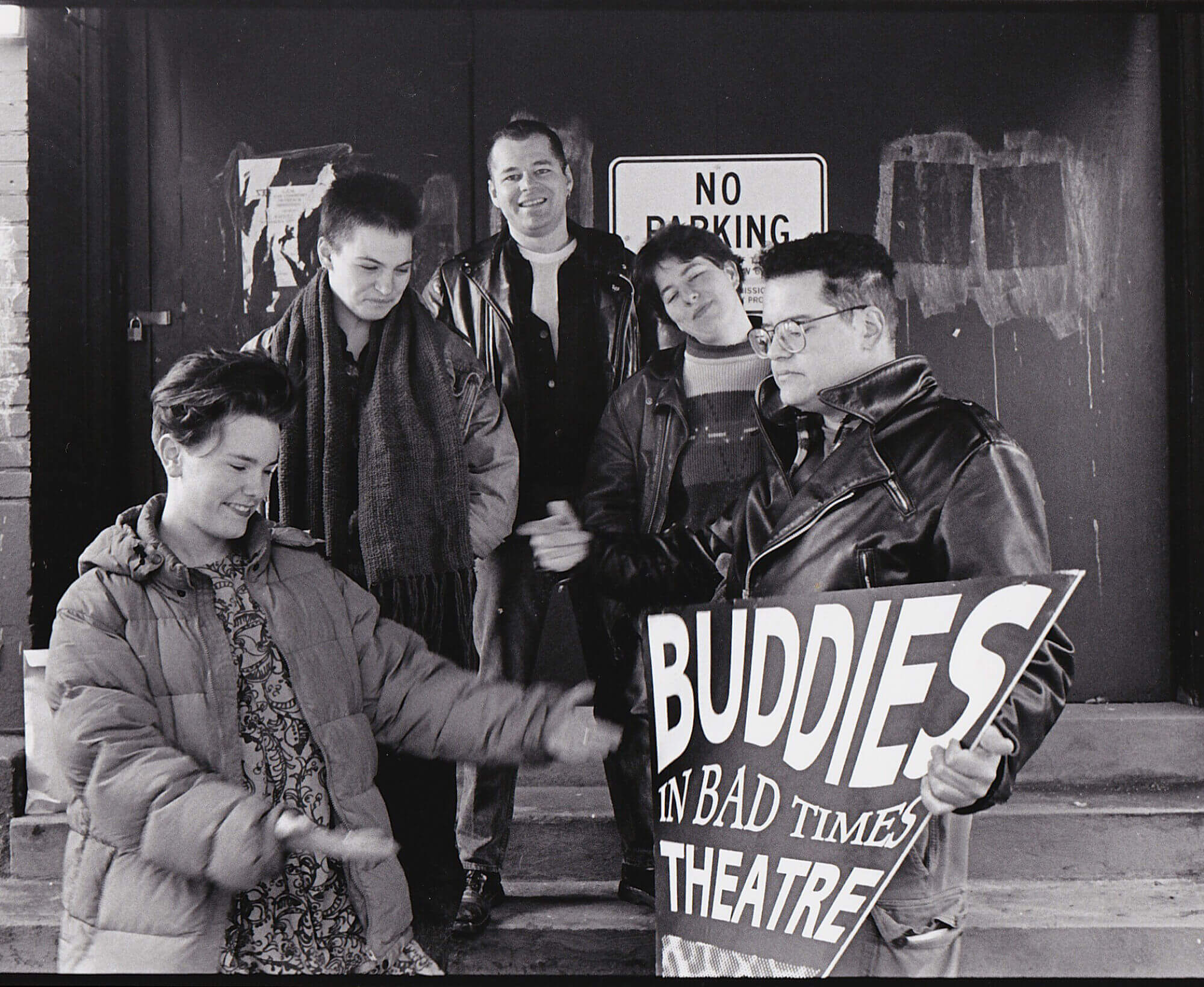 Black and white photograph of five people standing in front of a theatre dressed in warm clothing holding a sign reading "Buddies in Bad Times Theatre"