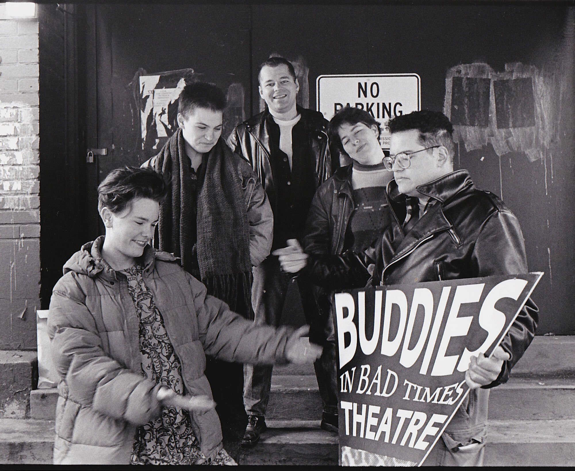 Black and white photograph 5 people standing in front of a theatre dressed in warm clothing holding a sign reading 