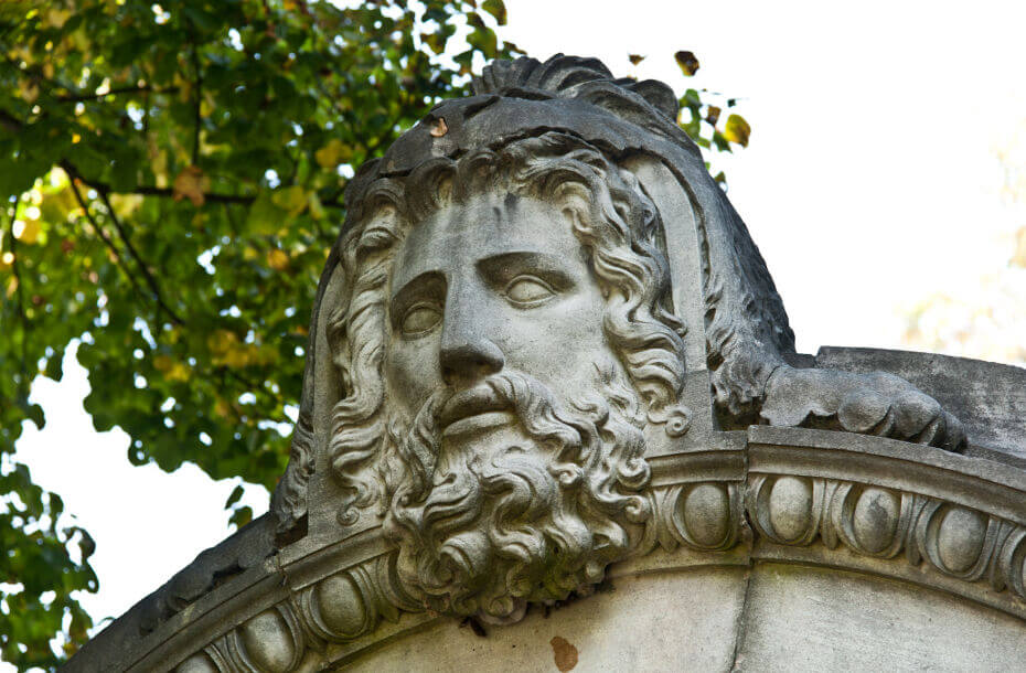 A carved stone face in front of a leafy background and sky.