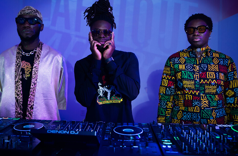 Three men stand behind a dj decks facing the camera. They are inside with blue lighting behind them. All three wear sunglasses.