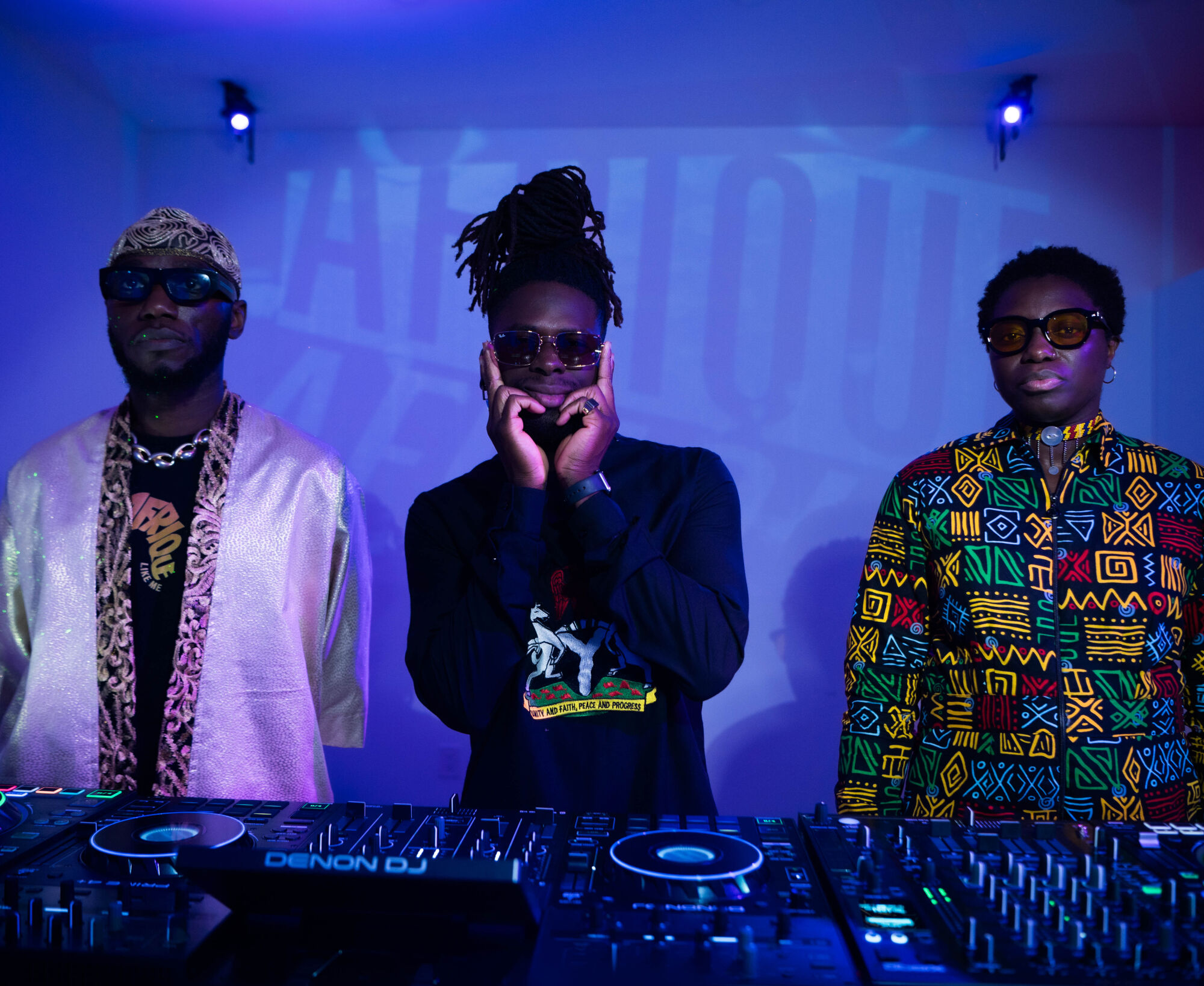 Three men stand behind a dj decks facing the camera. They are inside with blue lighting behind them. All three wear sunglasses.