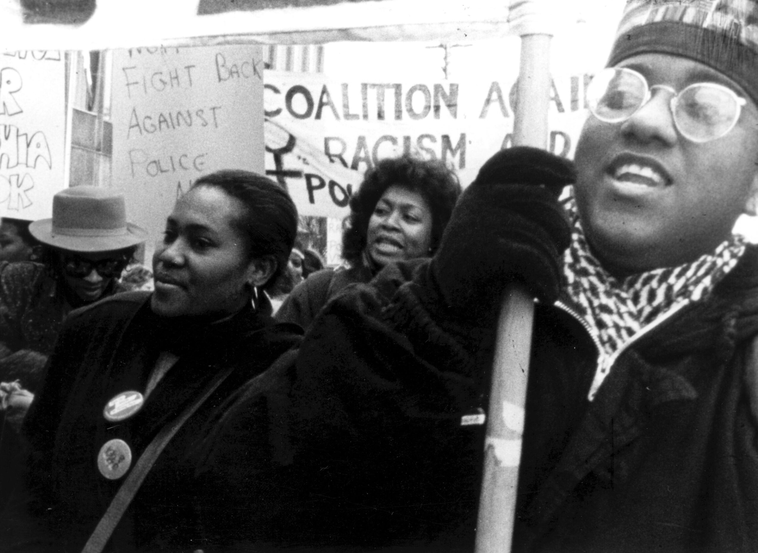 A black and white image of a group of women protesting with signs that say "fight back against police" and a cutoff sign that has the words "coalition and racism"