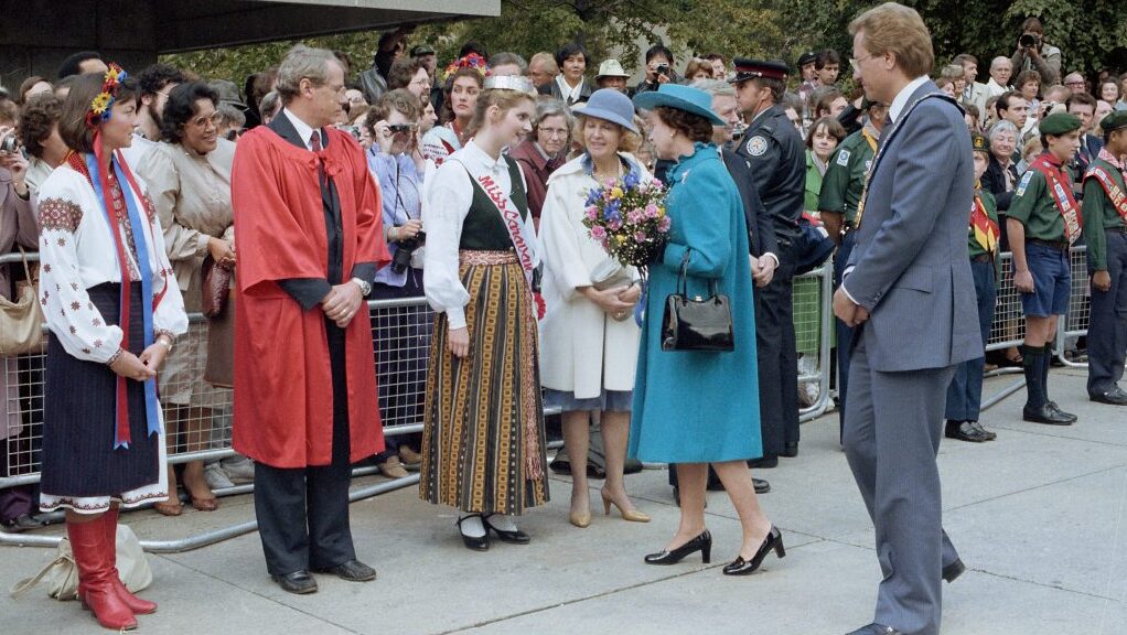 Queen Elizabeth II and Mayor Art Eggleton are greeted by a large crowd at the Metro Toronto International Caravan. The Queen speaks directly to the Miss Caravan representative, and several other representatives of the festival.