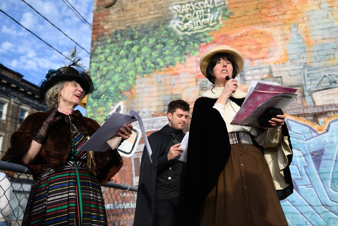 Three people stand in front of a colourful painted brick wall. They all are wearing typical nineteenth-century clothing. The woman on the right holds a microphone and papers in her hands.