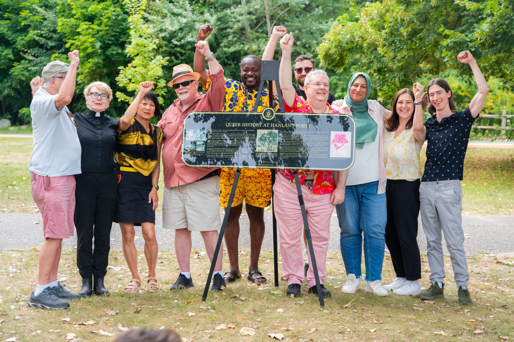 Toronto Mayor Olivia Chow, Deputy Mayor Ausma Malik, and members of the group Friends of Hanlan's stand with smiles and fists raised in a group photo on the grass in the park behind a new heritage plaque.