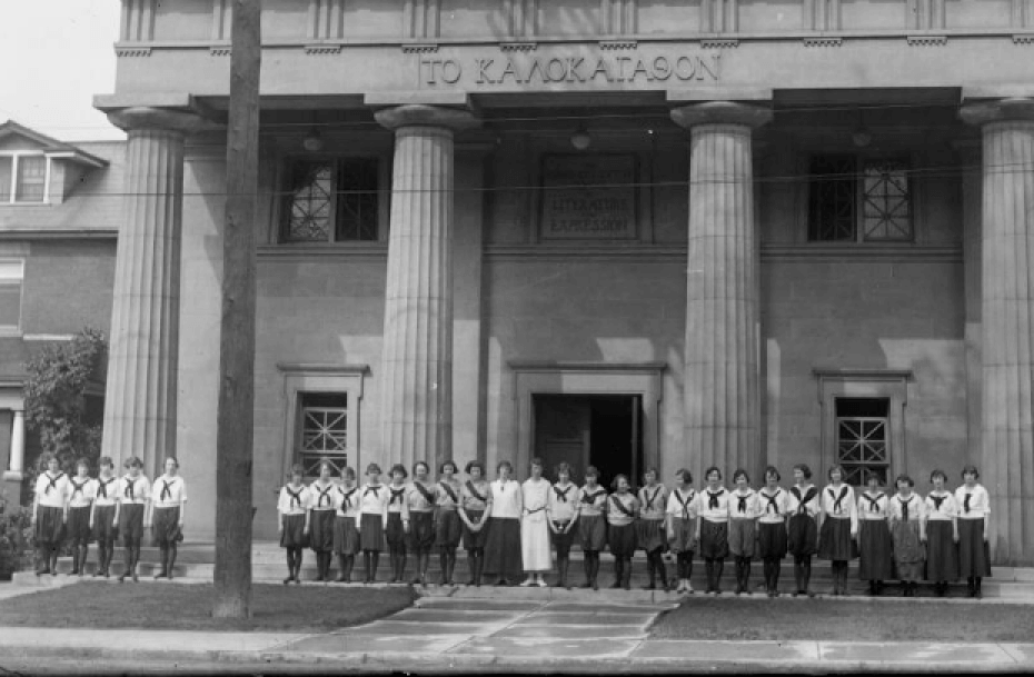 An archival photo shows a line of women in uniforms consisting of dark leggings, knee or ankle length skirts and neckerchiefs. They are standing in front of a building with four large columns that are two stories high.
