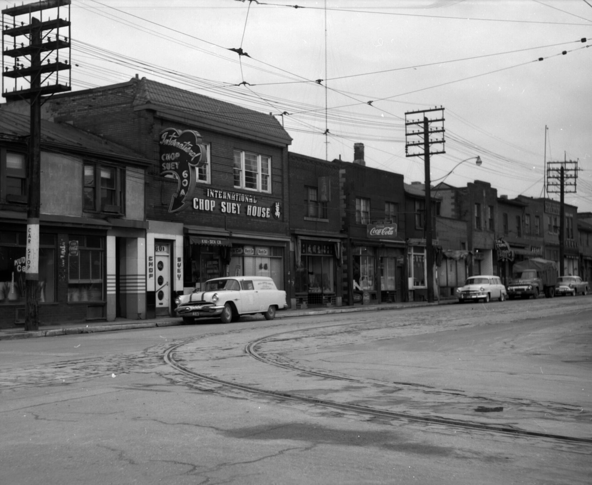 A black and white photograph depicting a street scene. A series of one and two-storey buildings can be seen behind a row of electric poles. The largest building has a sign out front that reads 