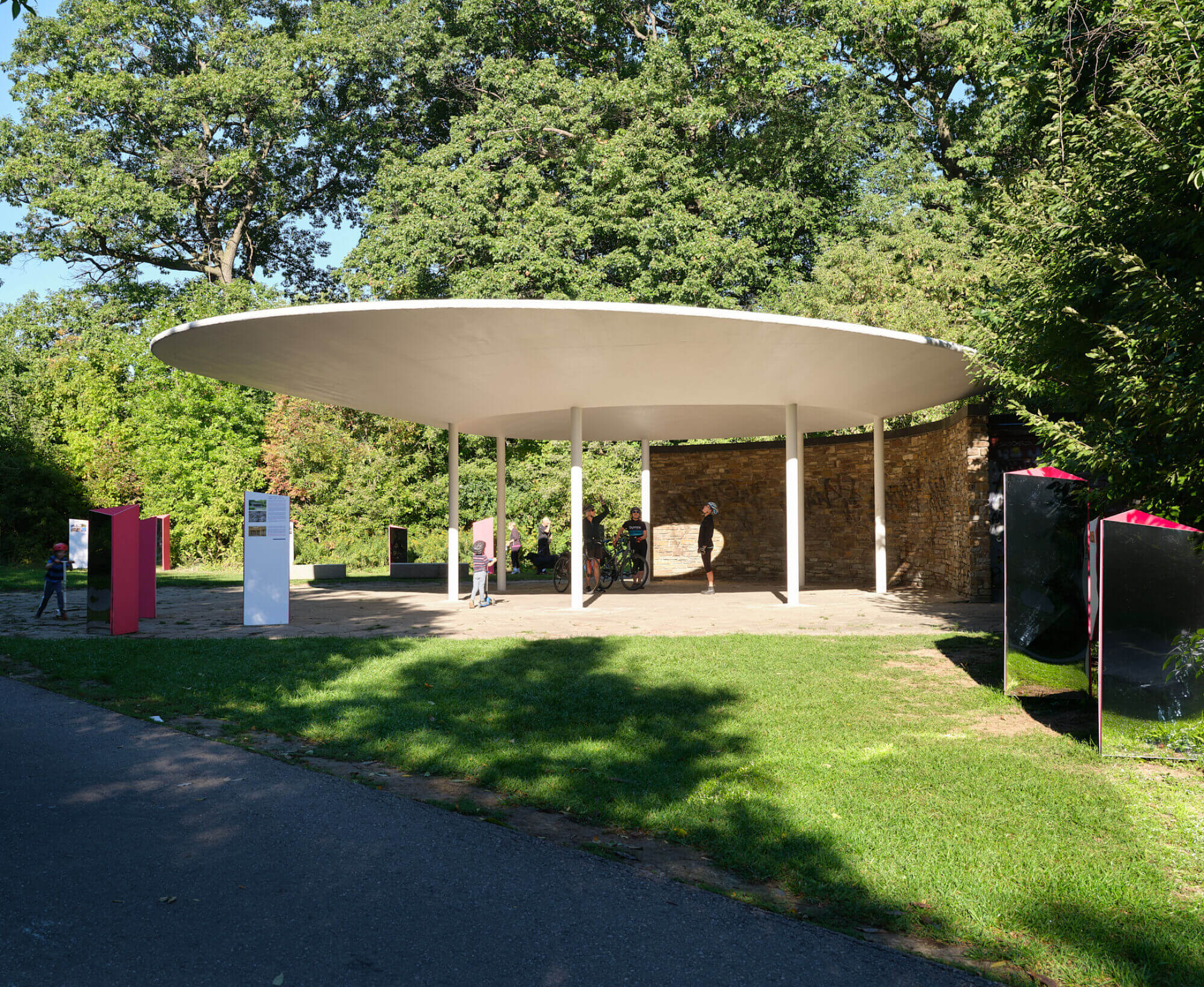 A large circular structure in a park. The structure is held up by polls. Behind the structure is a curved brick wall. A variety of heritage plaques and reflective posts cover the space around the structure.