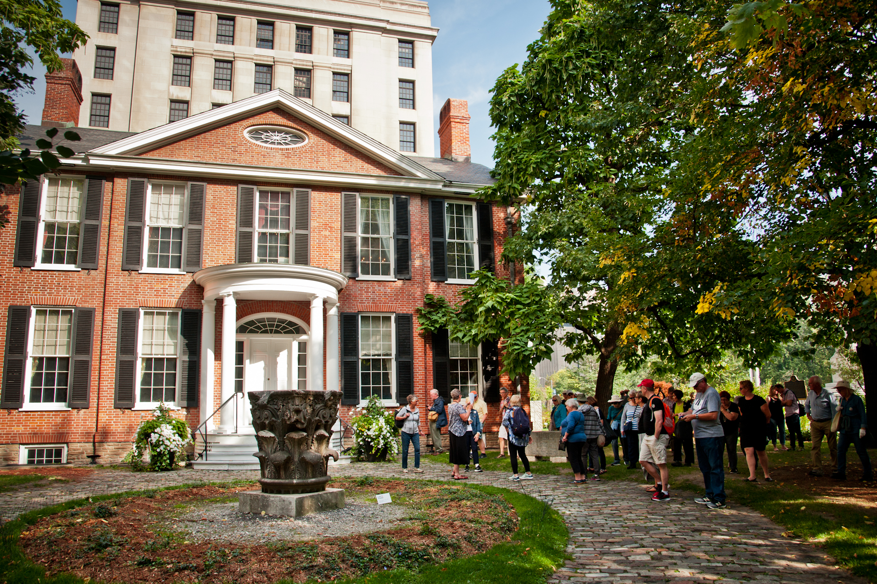 Old brick building with many rectangle windows in bright sunlight with a group of people looking at it.
