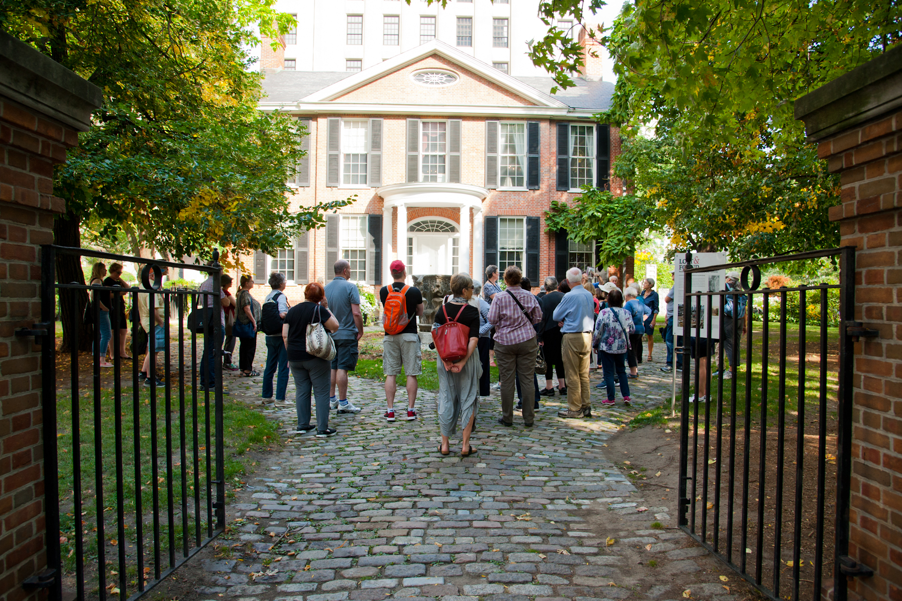 A group of people looking at a building can be viewed from behind, through a wrought iron gate. The building is the Campbell House Museum.
