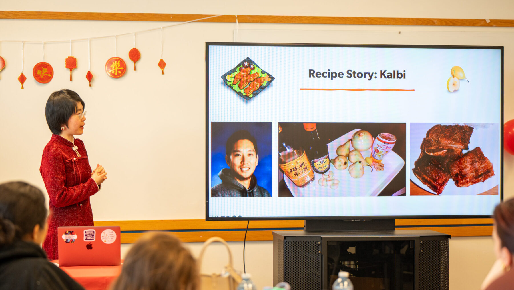 Person stands at the left and looks to the right where a presentation screen is visible with the title Recipe Story: Kalbi with images and illustrations. The back of peoples' heads are seen in the foreground.