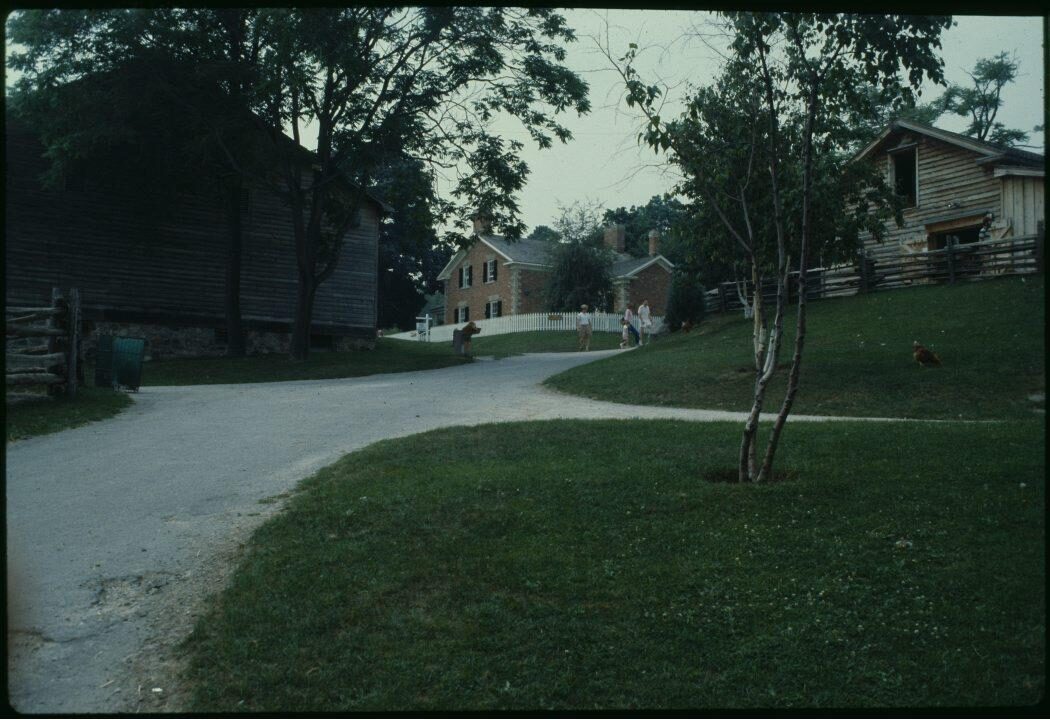 Several farm buildings in a park-like environment, connected by gravel paths. The buildings consist of two barns and a Victorian-style farmhouse. In the background, several people are walking along the paths.