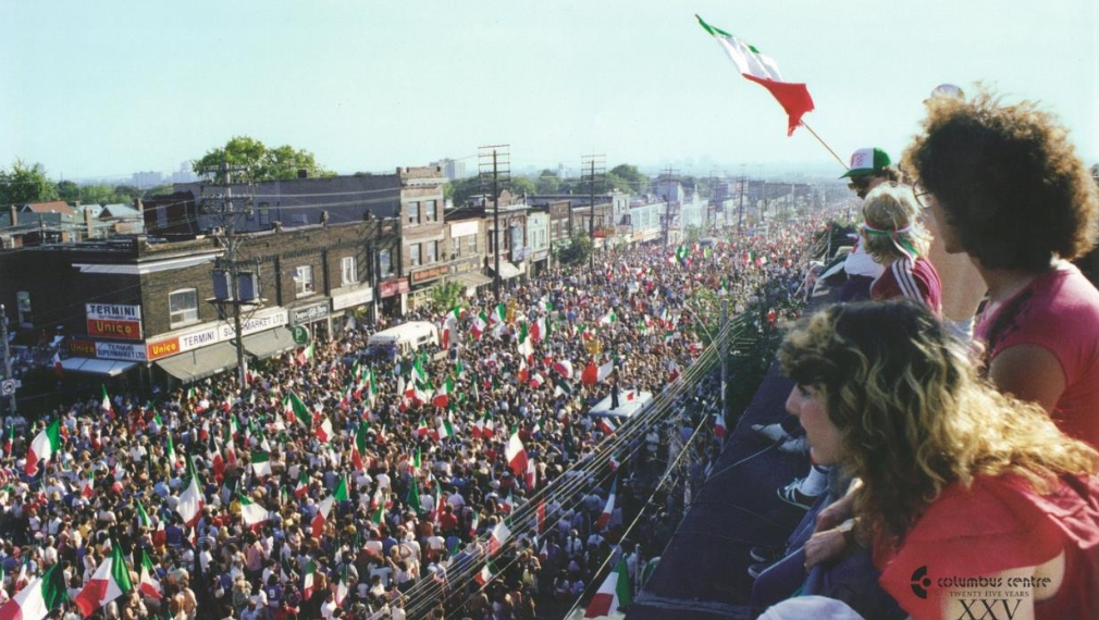 Two people in the foreground looking down to an extremely crowded street of people celebrating and waving the flag of Italy.