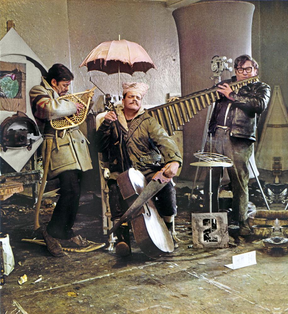 Members of a band posing humorously with pretend instruments. One man sits playing a broken cello holding a hand saw. To his right, a man stands, posing with a snowshoe for a guitar. On the right, another man holds organ pipes to mimic playing the flute.