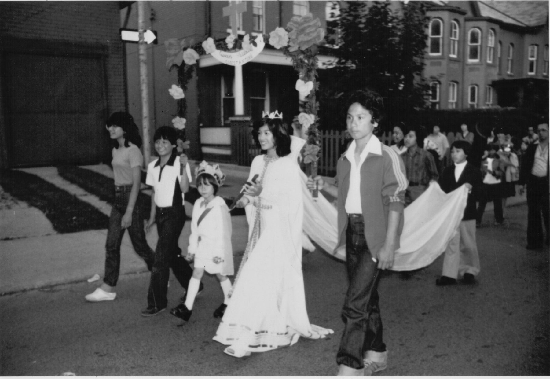 A black and white photograph of a Filipino Woman in a white dress walking as part of a procession for a Filipino ceremony, flanked by a man and three children, with a child carrying the train of her dress behind her.