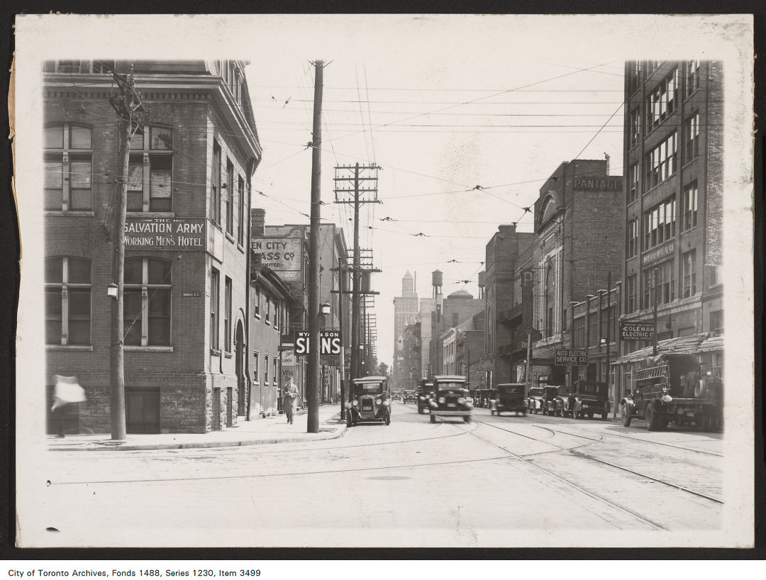 A black and white photograph of a street scene. Several brick buildings on either side of the road. Old cars from the 1920s can also be seen driving and parked on the road.